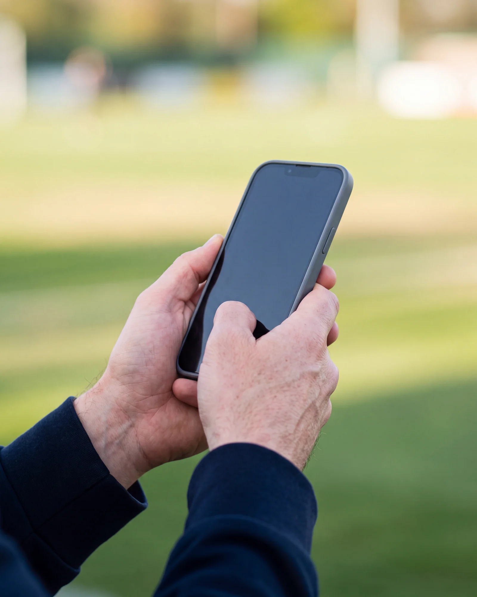 グラウンドを背にして、両手でスマートフォンを持っている手元。画面は暗いまま。
